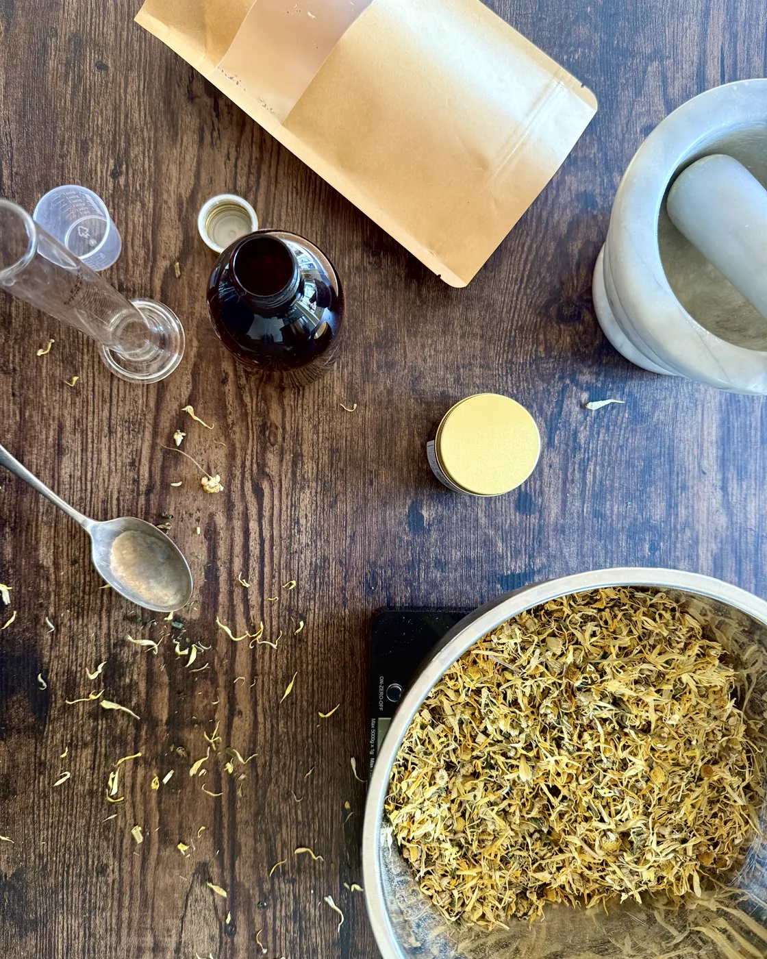 Dried calendula flowers in a bowl with a pestle and mortar and an amber tincture bottle on a wooden bench